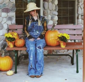 image of a scare crow costumed woman sitting on a porch bench surrounded by pumpkins